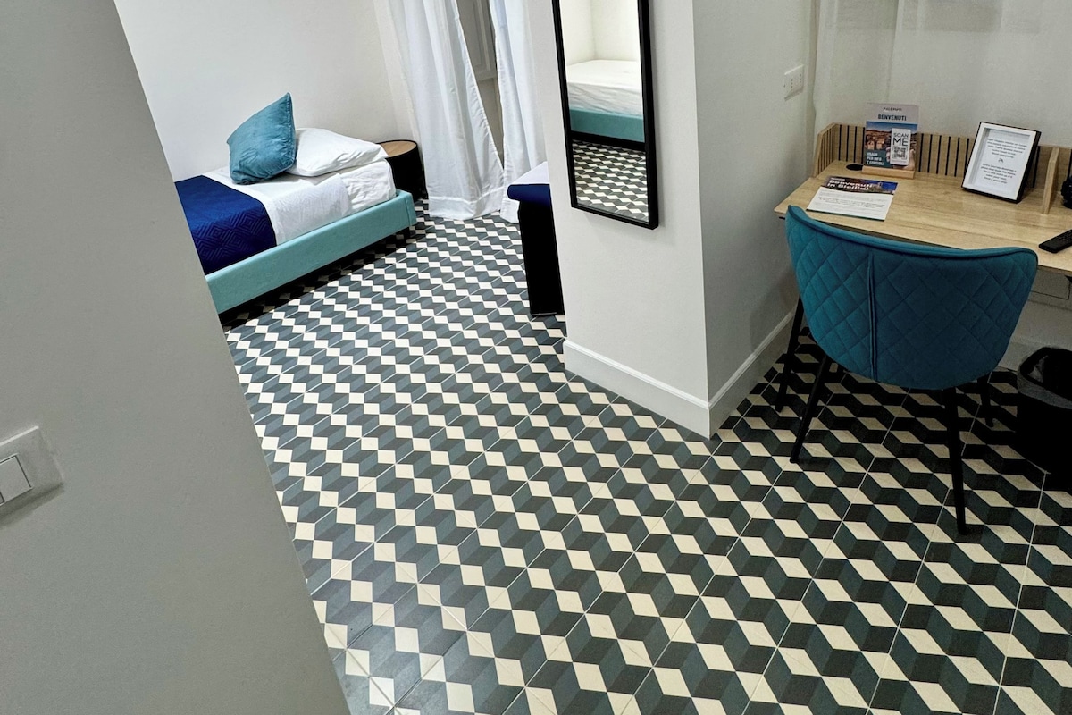 Una habitación con piso a cuadros, una cama con ropa de cama azul, un escritorio con una silla y un espejo en la pared.
