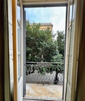 Vista da un balcone con ringhiera in metallo, un edificio con facciata gialla e alberi sullo sfondo.