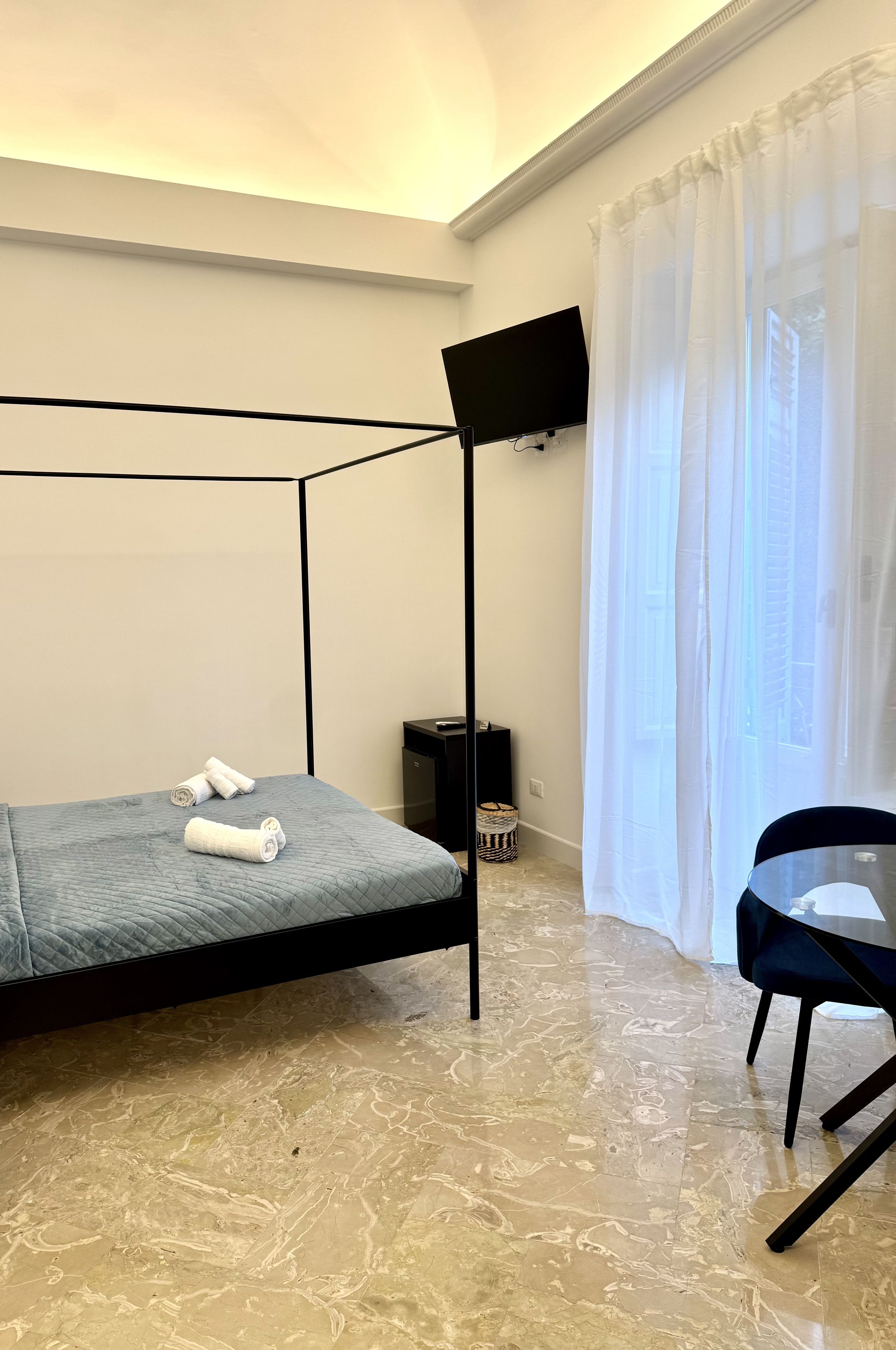 Ein Zimmer mit einem Bett, einem an der Wand befestigten Fernseher, einem Sessel und einem Fenster mit Vorhängen.