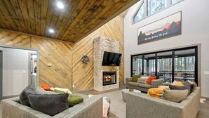A modern living room with a stone fireplace, grey sofas, and a large window overlooking a forested area.