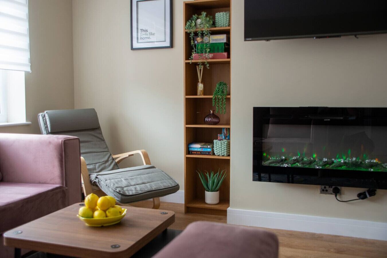 Een woonkamer met een bank, een fauteuil, een bijzettafeltje met citroenen, een boekenkast met planten en een aan de muur bevestigde televisie.