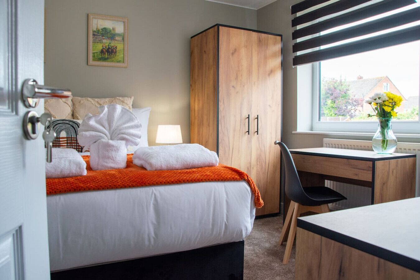 A hotel room with a bed, a wooden wardrobe, a desk, and a window with a view of houses.