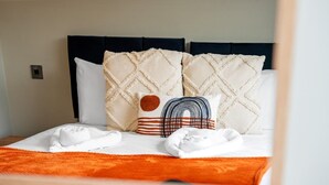 Een keurig opgemaakt bed met een witte deken, twee kussens en een oranje plaid.