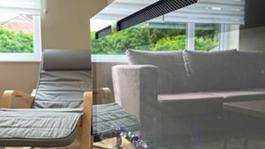 Een kamer met een houten vloer, een grijze bank, een fauteuil en een groot raam met zonwering.
