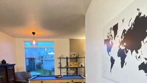A living room with a pool table, a white fluffy chair, a sofa, a world map on the wall, and a window with a view of a backyard. - 4-bedroom house in Höfn í Hornafirði (Höfn í Hornafirði)