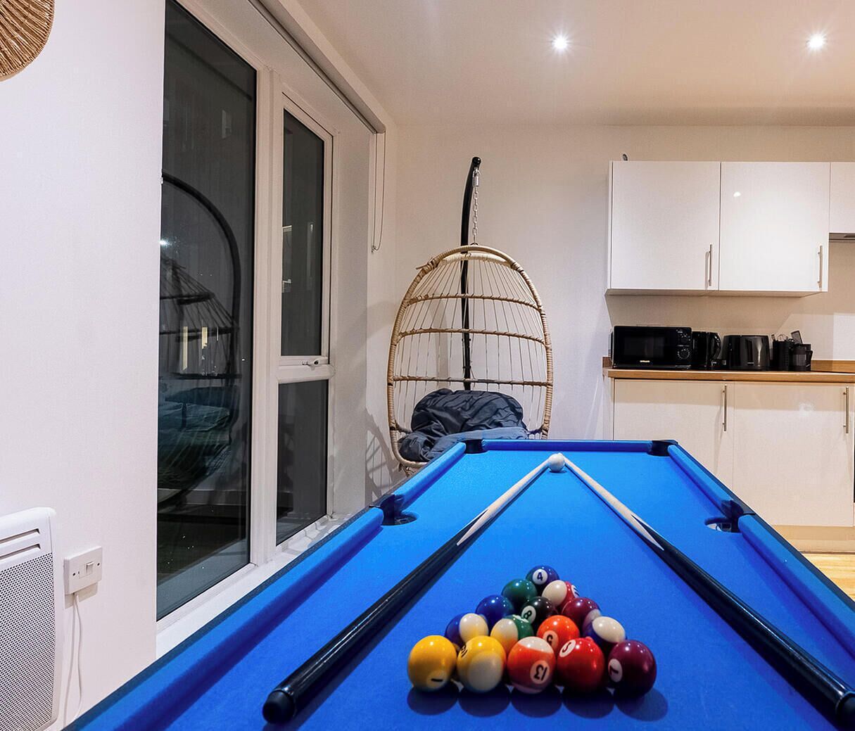 A pool table with balls, a wicker chair, and a kitchen area with cabinets.