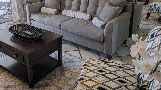 Una sala de estar con un sofá gris, una mesa de centro de madera oscura, una alfombra estampada y un espejo grande que refleja la habitación.