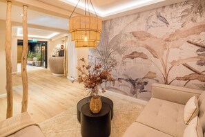 A modern lobby with a wooden floor, a black round table with a vase, a beige sofa, and a large wall mural. - Das Oswald's Boutique Hotel (Cuxhaven)