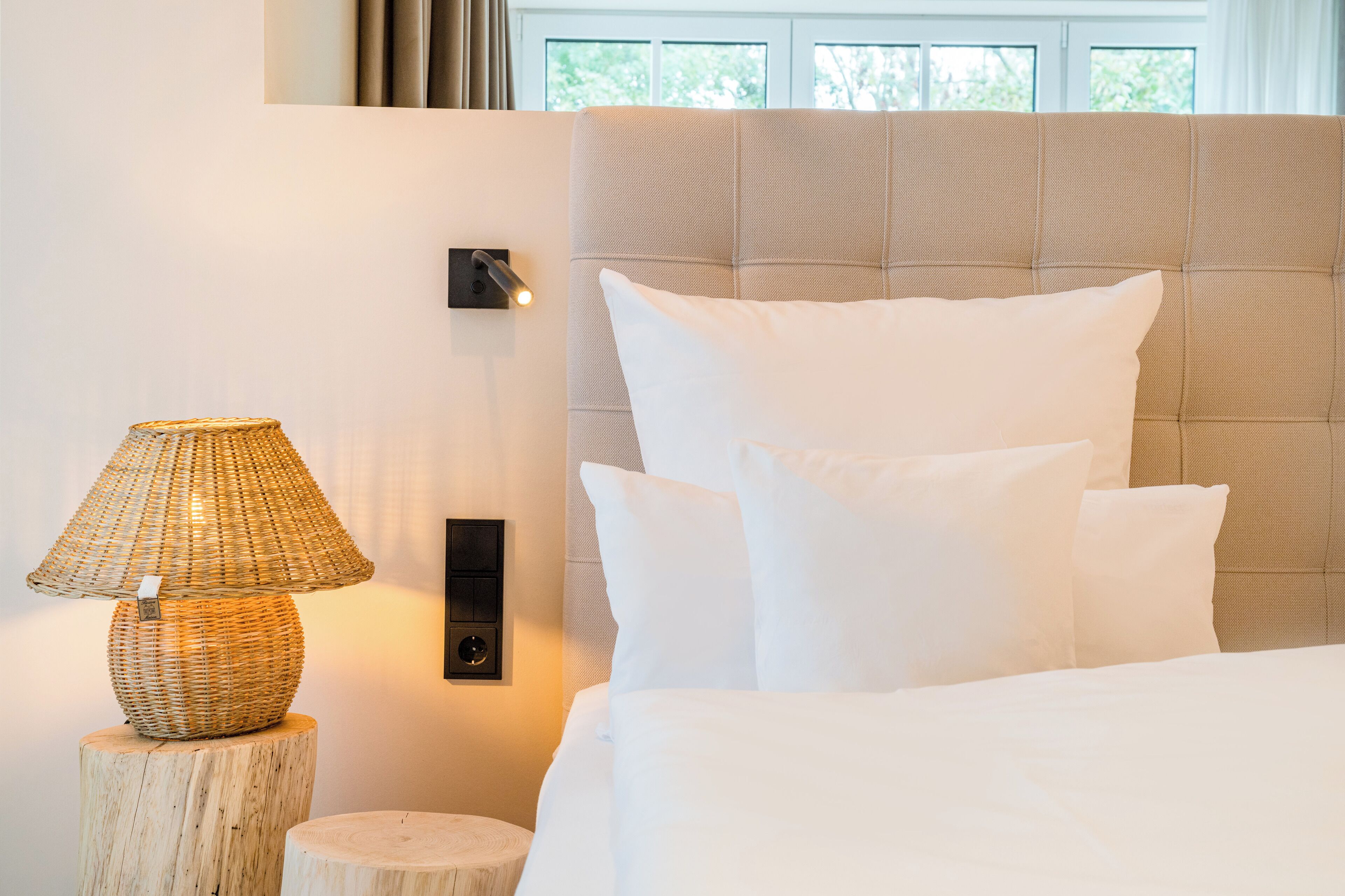 A neatly made bed with white bedding and a headboard, a wall-mounted lamp, and a wooden stool.