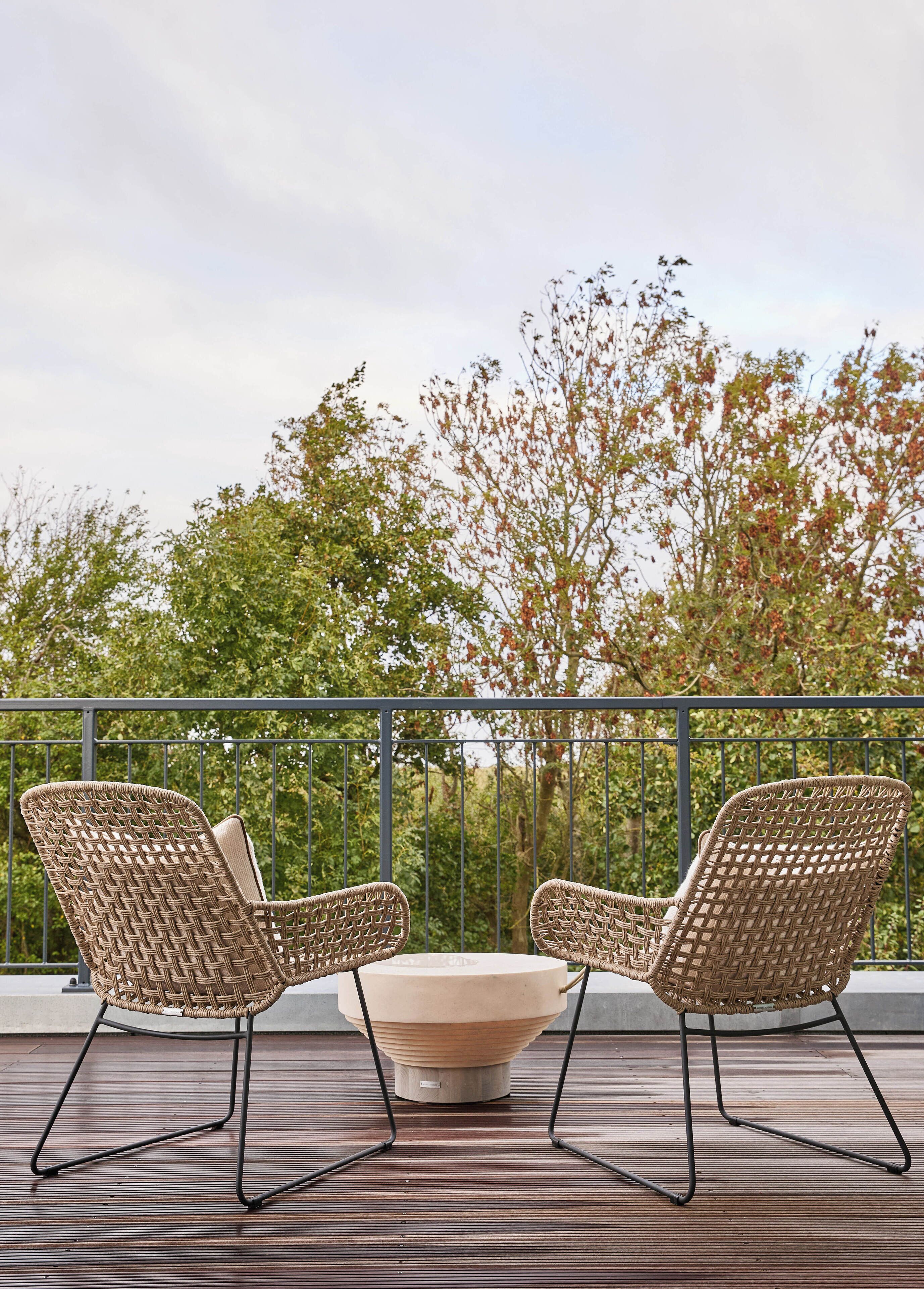 Two wicker chairs with metal legs and a central table on a wooden deck with a metal railing and trees in the background.