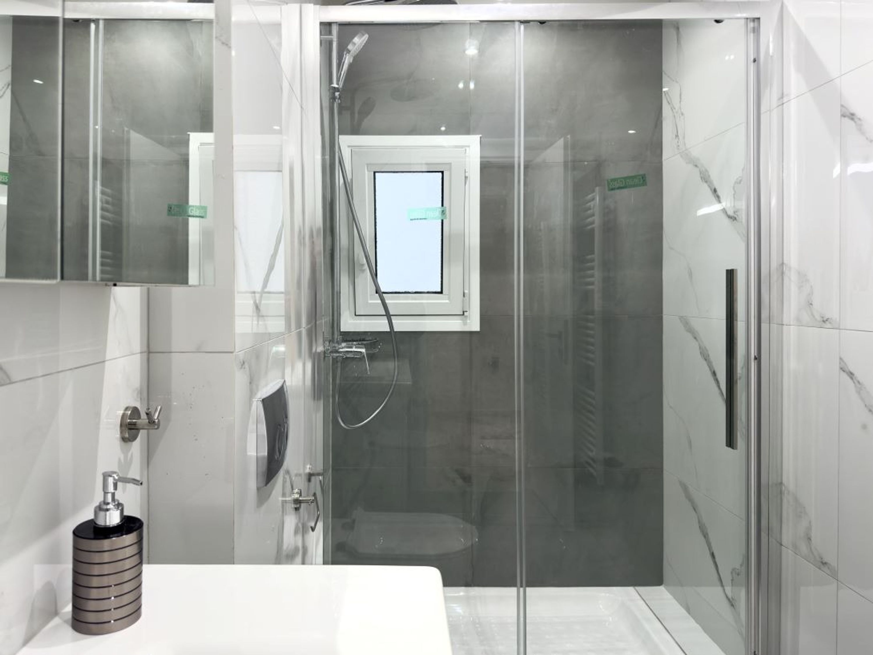 Un baño moderno con ducha envuelta en vidrio, un ventanal visible y un dispensador de jabón en el lavamanos.