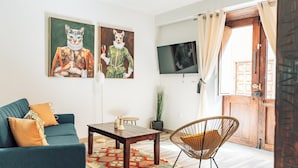 A living room with a sofa, coffee table, and a chair. There are two paintings on the wall and a TV mounted above the door.