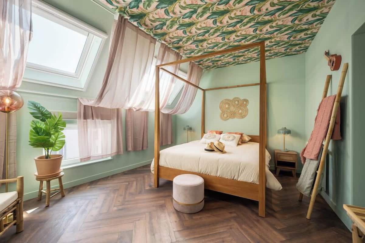 A four-poster bed with a wooden frame and a round ottoman in front.