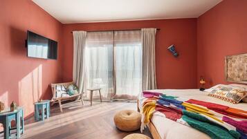 A bedroom with a bed, a TV, a chair, a small table, and a window with curtains.