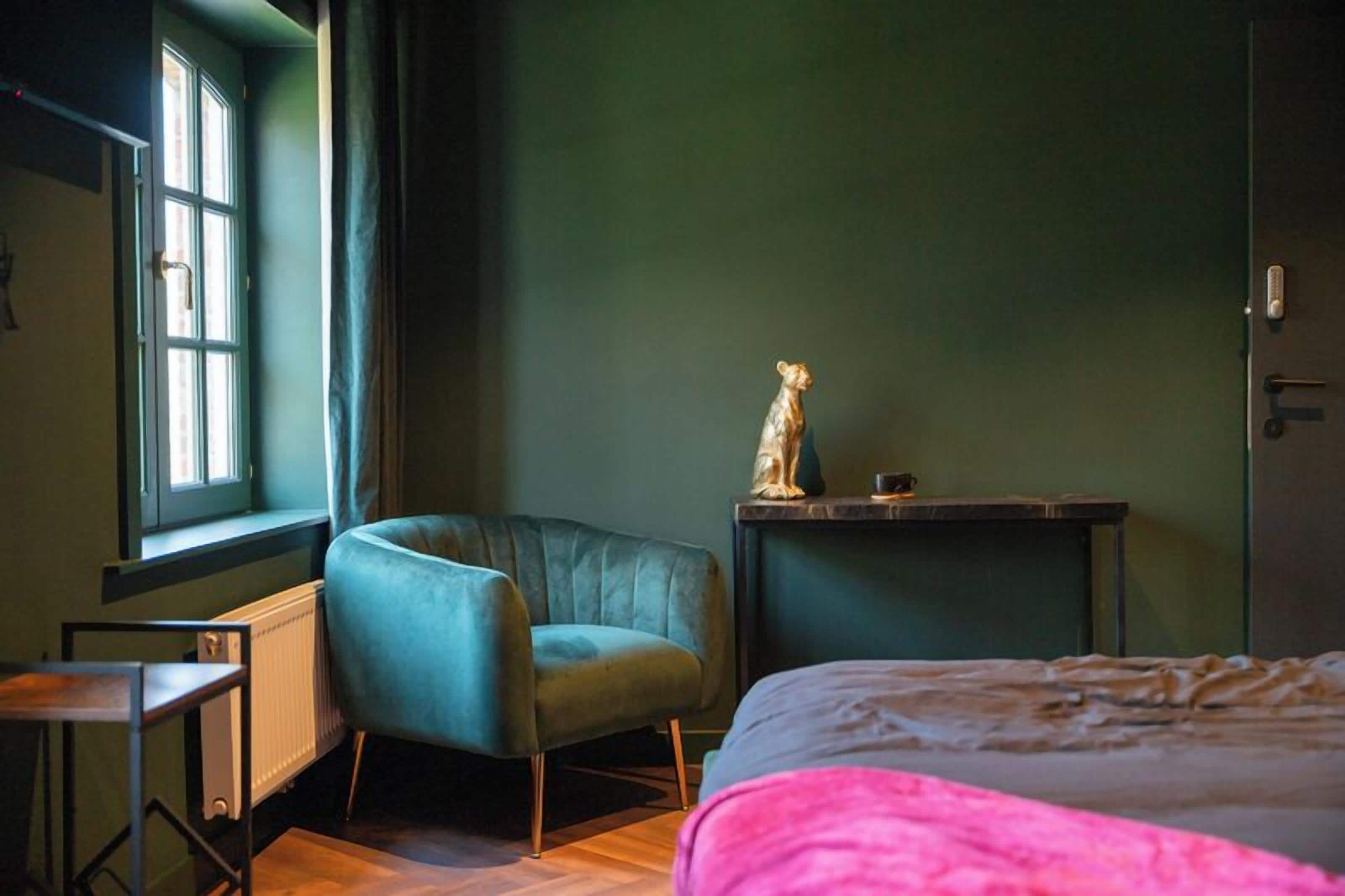 A room with a teal armchair, a bed with a pink blanket, a small table with a statue, and a window with curtains.