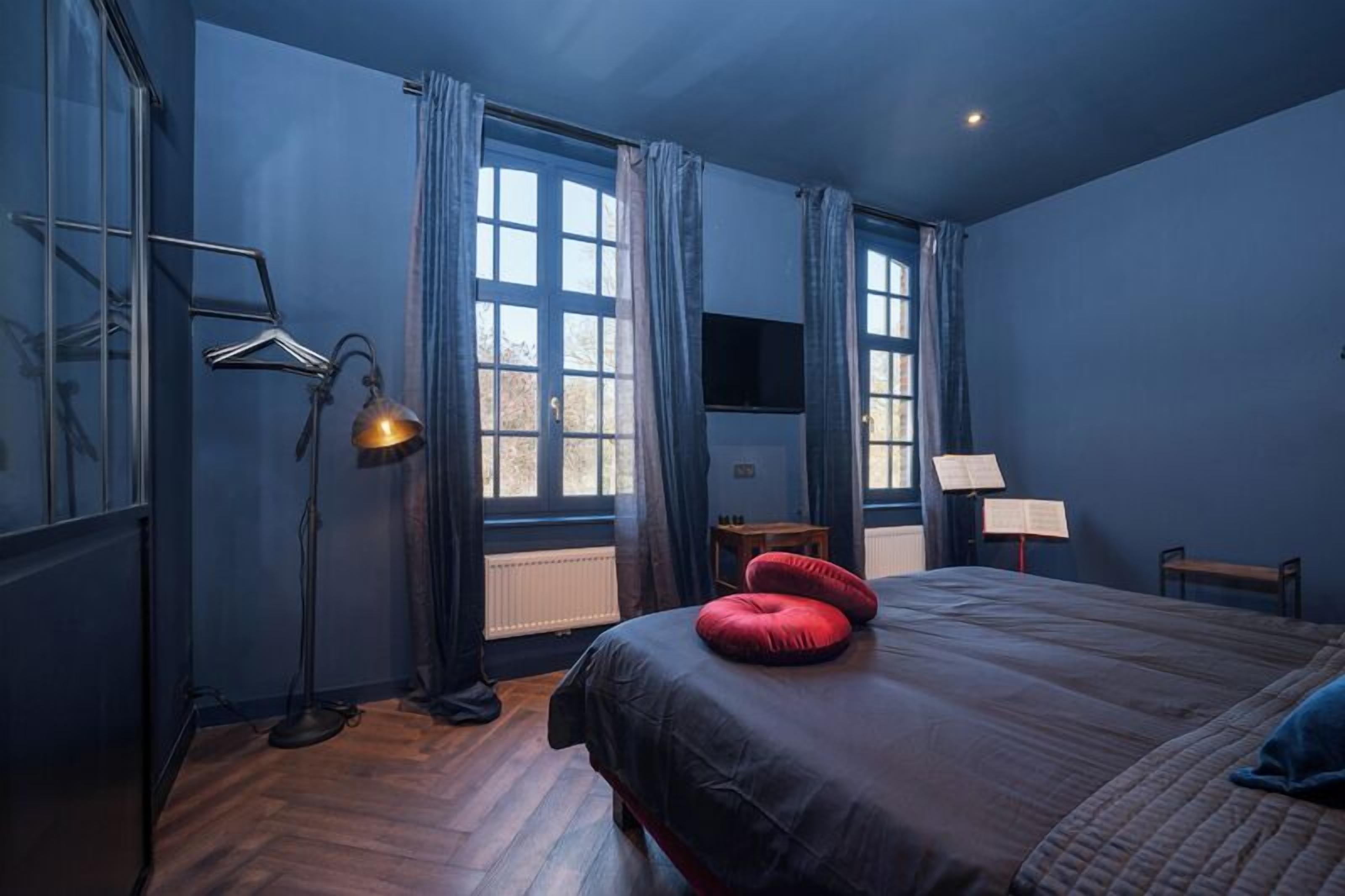 Un dormitorio con una cama grande, un escritorio con silla, una lámpara de pie y una ventana con cortinas.