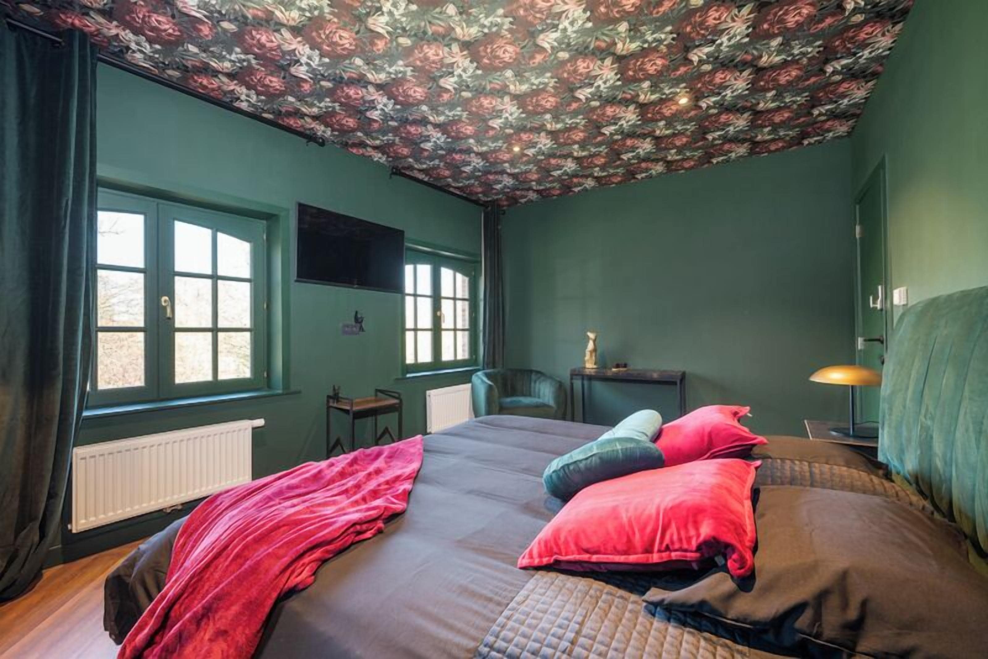 A bedroom with a floral ceiling, a bed with multiple pillows, a window, a radiator, a television on the wall, and a lamp on a bedside table.