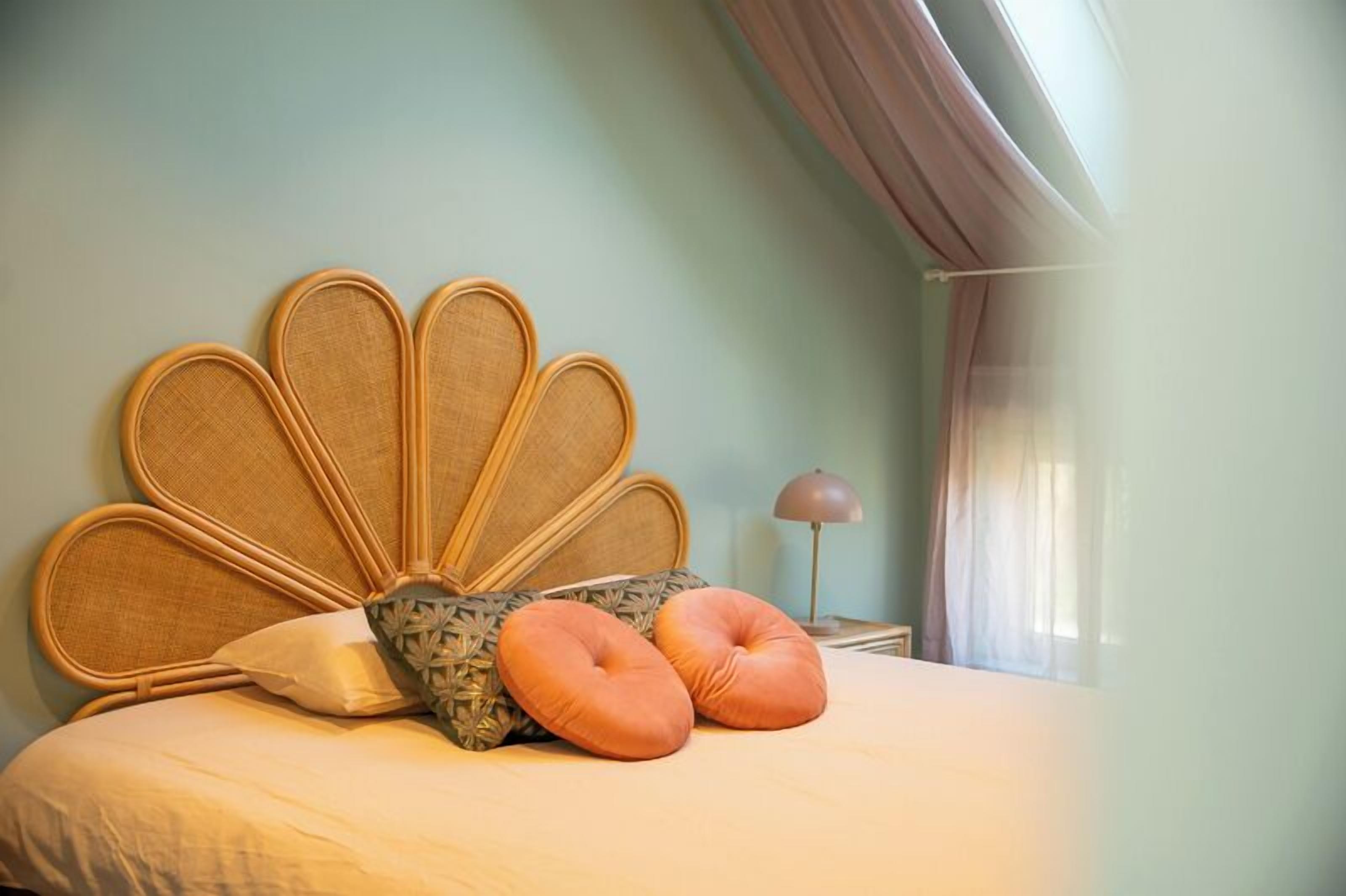 Cama com cabeceira de vime, duas almofadas laranja e uma almofada estampada.