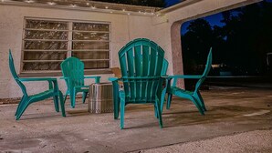 An outdoor seating area with green chairs and a table, under string lights, with a building in the background.