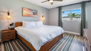 A neatly made bed with a wooden headboard, a ceiling fan, and a view of palm trees through a window.