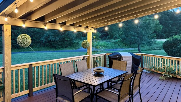 A wooden deck with a table and chairs, string lights, and a view of a landscaped garden. - Secluded, elegant country home only 10 minutes from Downtown Ithaca! (Ithaca)