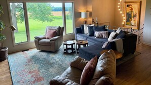 A living room with a large window, a sofa, a chair, a lamp, and a painting on the wall. - Secluded, elegant country home only 10 minutes from Downtown Ithaca! (Ithaca)