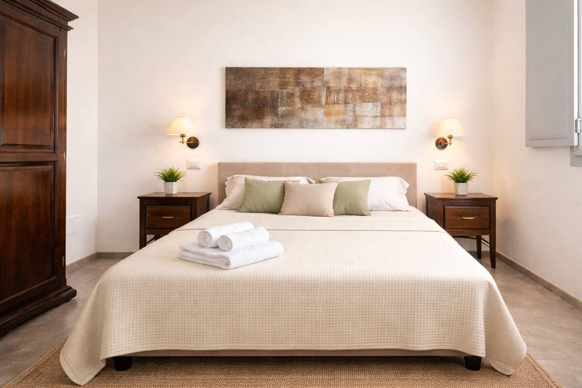 A neatly made bed with a beige bedspread, two bedside tables with lamps, a large abstract painting on the wall, and a wooden wardrobe.
