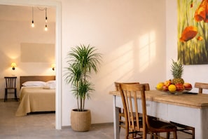 Ein Zimmer mit einem Bett, einem Esstisch mit Stühlen, einer Zimmerpflanze und einer Obstschale.