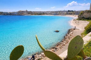A coastal view with clear blue waters, a rocky shoreline, and buildings in the distance. - Masseria Luci Grande (Scorrano)