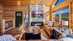 A cozy log cabin interior with a stone fireplace, a leather sofa, and a large window offering a mountain view. - Ridgetop Retreat - Epic Views - Hot Tub - Fire Pit - Kid Friendly - Arcade (Sevierville)