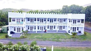 Un bâtiment blanc à plusieurs étages, doté de balcons et d’un toit plat, entouré de verdure.