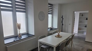 Moderni ruokailutila, jossa on valkoinen pöytä ja tuolit, seinällä oleva kello ja ikkunassa sälekaihtimet.