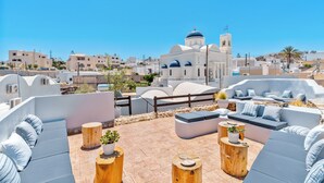 A rooftop terrace with outdoor seating, a view of a white church with a blue dome, and a clear blue sky. - ArtChroma House (Santorini)