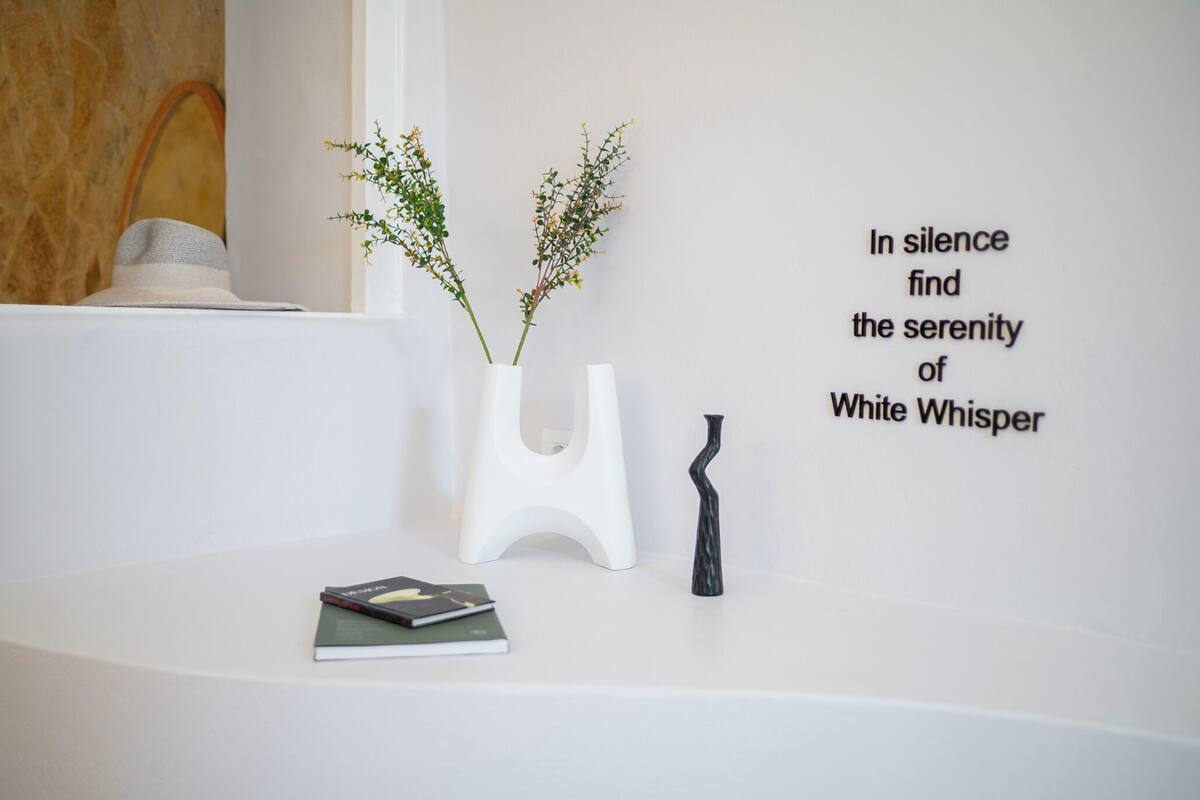 A white wall with a quote, a vase with plants, and a book on a white surface.
