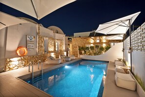 A pool area with stone walls, white furniture, and umbrellas. - ArtChroma House (Santorini)