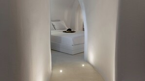 A narrow, white-walled hallway leading to a bedroom with a bed and a nightstand. - ArtChroma House (Santorini)