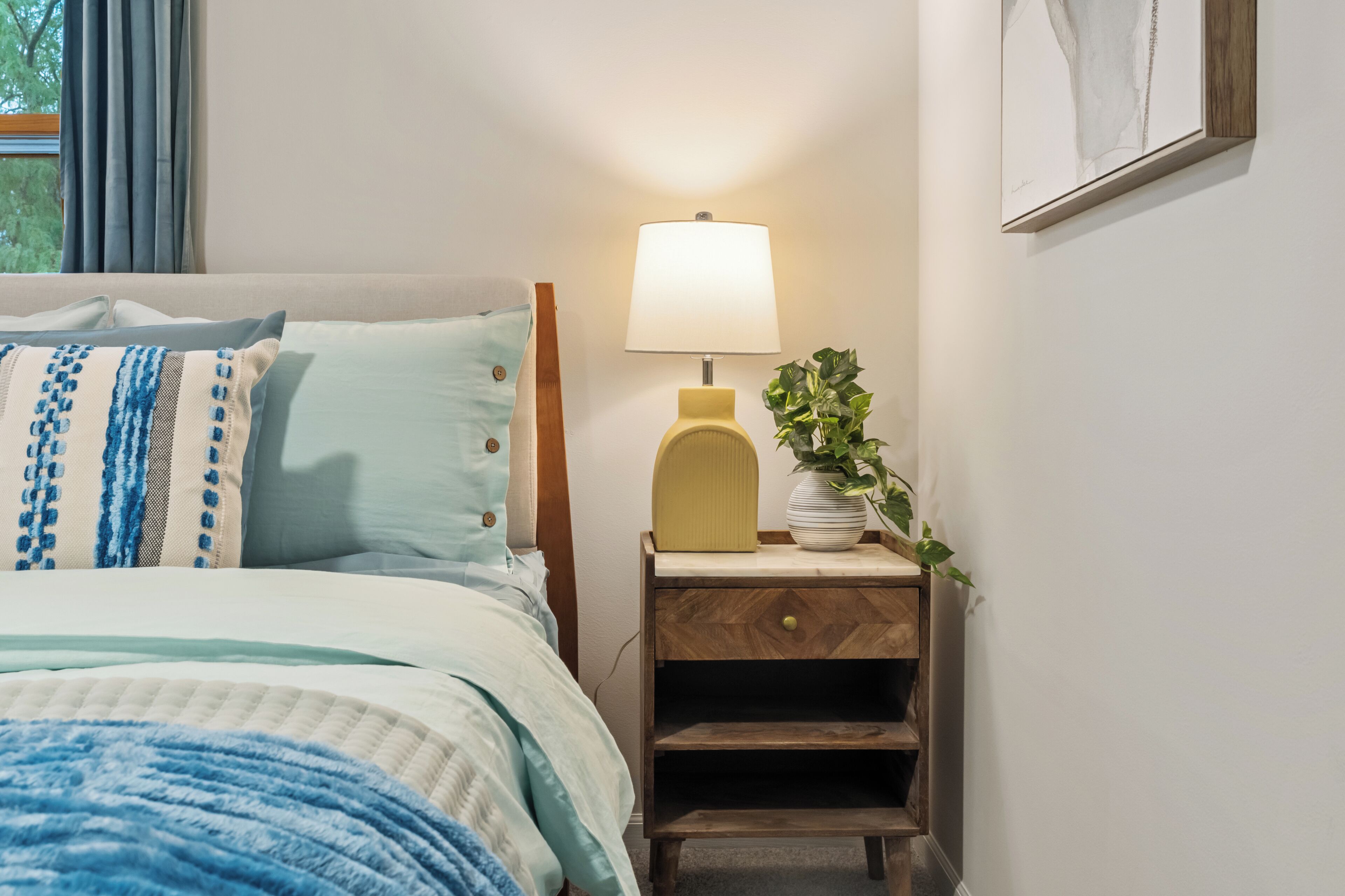 A neatly made bed with a blue and white striped pillow, a bedside lamp, and a small wooden nightstand with a plant.