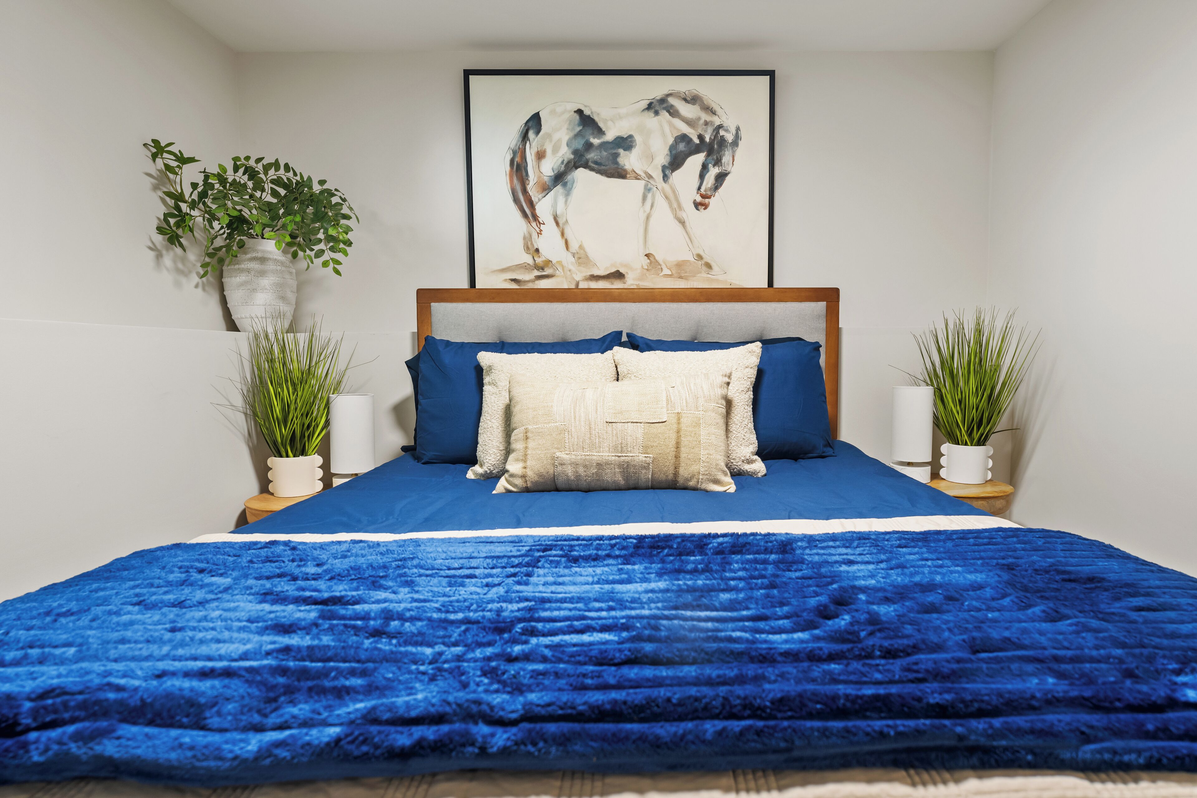 A bedroom with a blue bedspread, a framed horse painting on the wall, and two potted plants on either side of the bed.