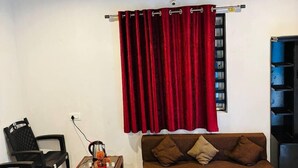 A living room with a brown sofa, a wooden coffee table, black chairs, a red curtain, and a ceiling fan. - COORG SUNRISE COTTAGES (Kushalnagar)