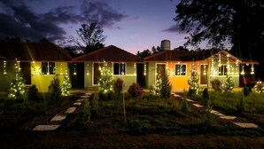 A two-story house with a well-lit exterior, surrounded by a garden with small trees and a stone pathway leading to the entrance. - COORG SUNRISE COTTAGES (Kushalnagar)