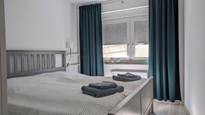 A neatly made bed with a grey headboard, a bedside lamp, and a window with curtains. - Hedel vacation apartment (Halle (Saale))