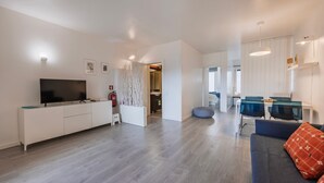 A modern living room with a flat-screen TV, white cabinetry, a dining area with blue chairs, and a sofa with a patterned cushion.