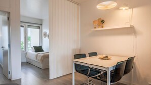 A modern, minimalistic room with a white table and chairs, a bed with a green pillow, and a window with blinds.