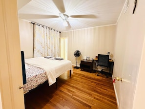 Un dormitorio con cama, escritorio, silla, ventilador de pie y ventilador de techo.