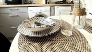 A modern kitchen with grey cabinets, a flat-screen TV, and a dining area with a woven placemat, a glass, and a vase with a plant. - Studio Lumière du Centre - Parking - Romans (Romans-sur-Isère)