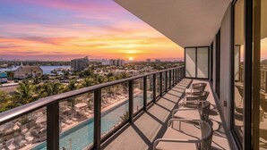 A balcony with a view of a sunset over a cityscape, a swimming pool, and lounge chairs. - Elegant Miami Beach Condos (Miami Beach)