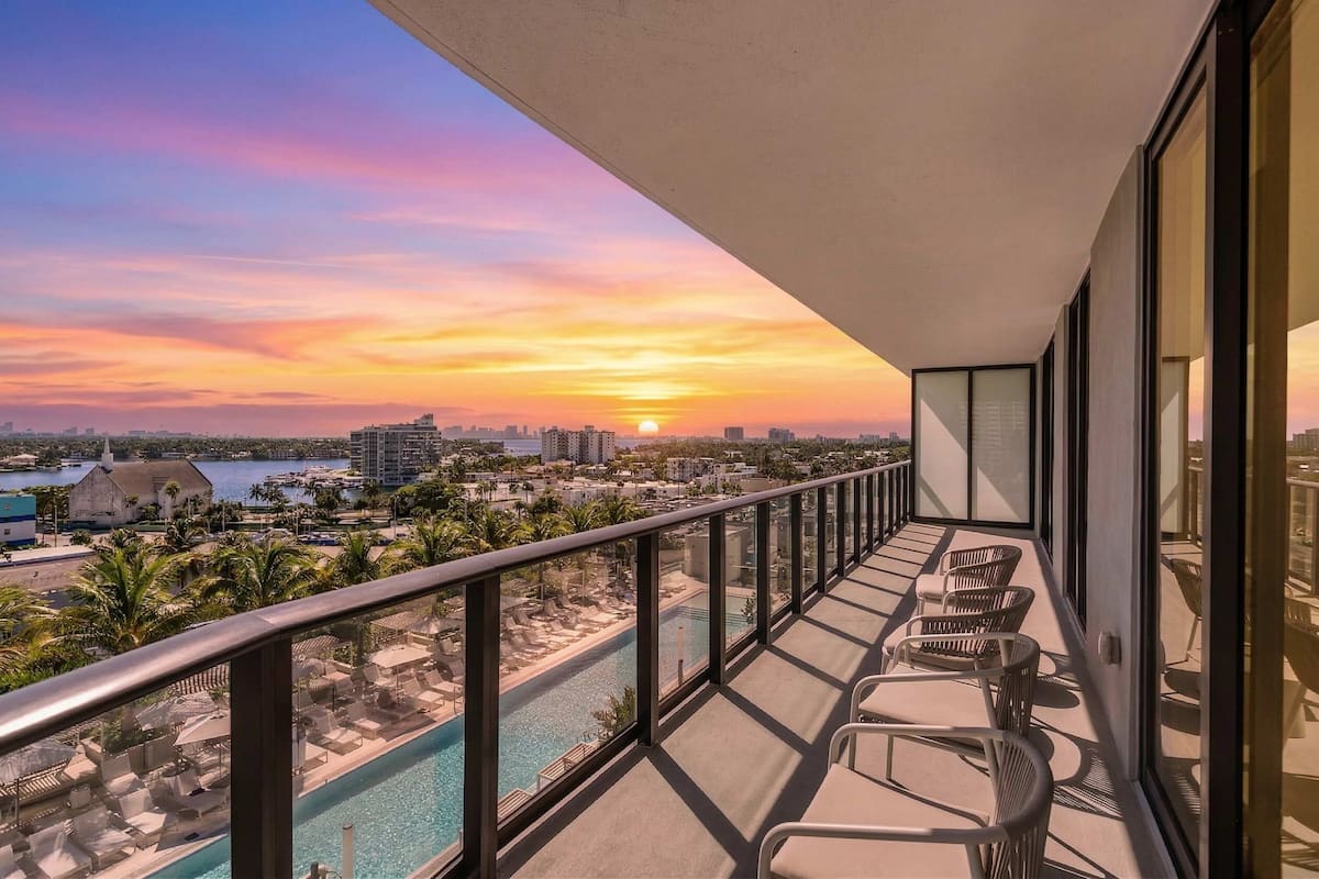 A balcony with a view of a sunset over a cityscape, a swimming pool, and lounge chairs.