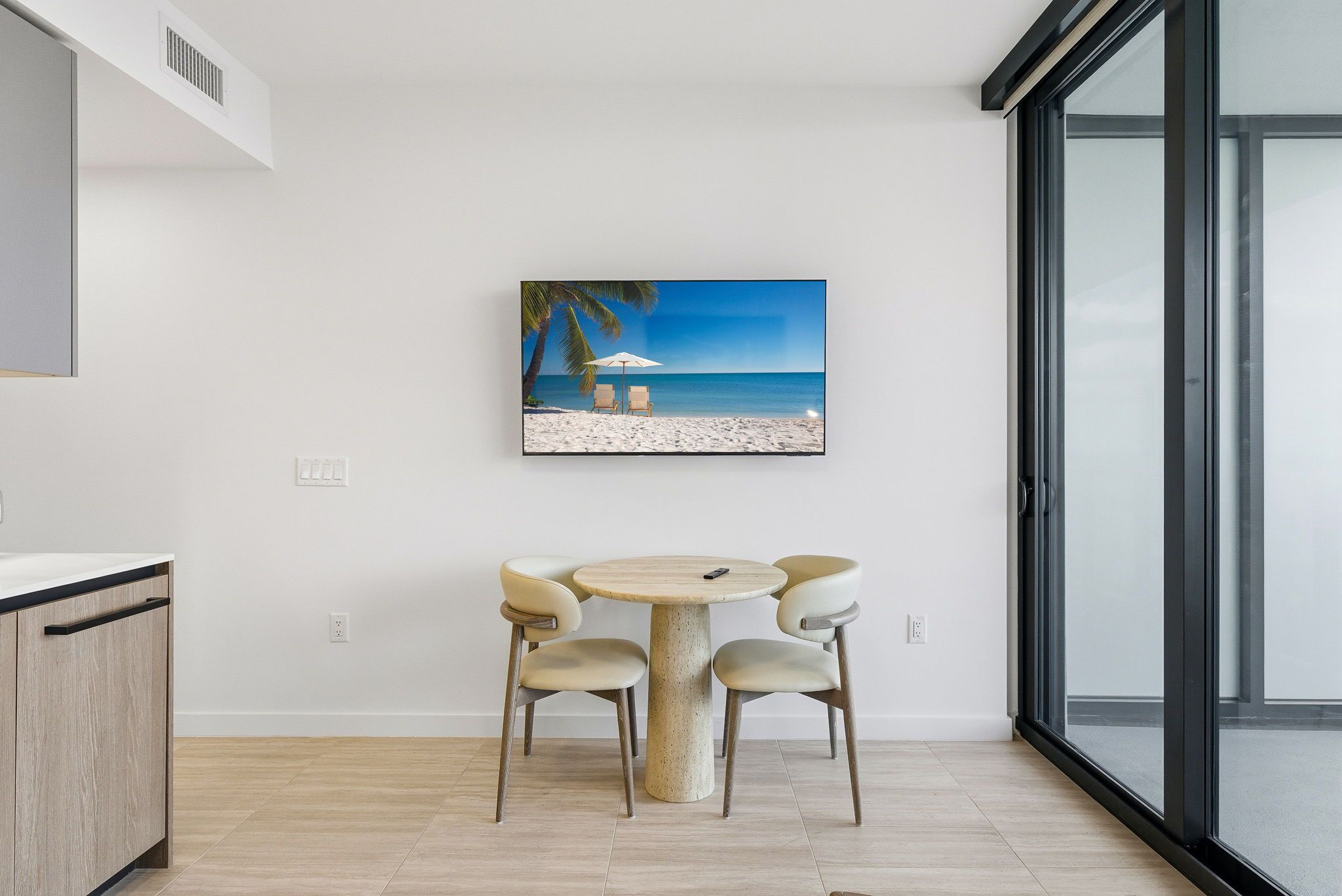 A dining area with a round table and four chairs, a painting of a beach scene, and a large window offering a view of the beach.