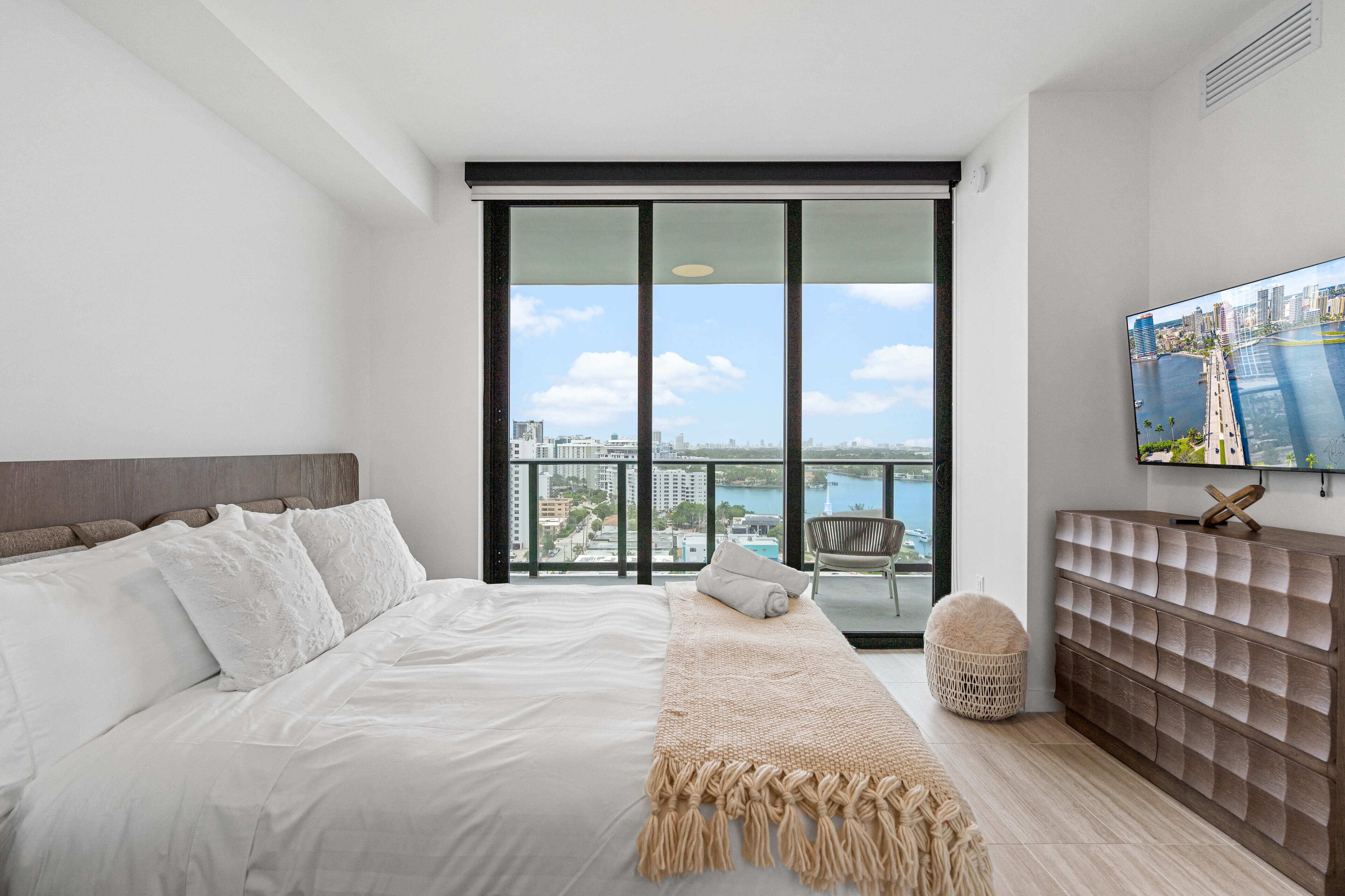 A modern bedroom with a large bed, a view of the city through floor-to-ceiling windows, and a mounted TV on the wall.
