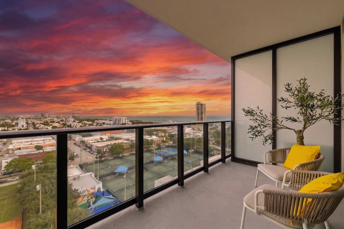 A balcony with a view of a cityscape during sunset, wicker chairs with yellow cushions, and a potted plant.